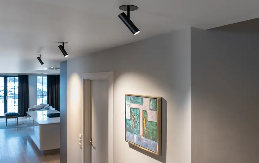Drei schwarze Aufbaustrahler an der Decke strahlen ein Bild an der Wand an
